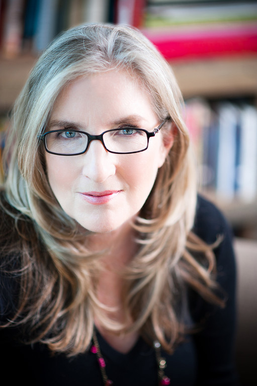 A photo of Luanne Rice. She is looking directly at the viewer with her blue eyes behind dark rimmed glasses. Her blonde hair comes down past her shoulders and she is smiling warmly. Behind her is an out of focus bookshelf full of books.