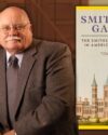 Author Tom Crouch with a white mustache, wearing thin framed glasses in a brown suit jacket, white button up shirt and brown tie, smiling. Features the book cover of Smithson's Gamble by Tom Crouch.
