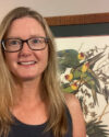 Presenter Jillian Liner smiling, wearing dark rimmed glasses. She has long blonde hair and a dark tanktop. Behind her is a framed print hanging on the wall featuring birds with a red and yellow head and and various shades of green on the body.