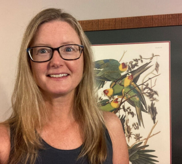 Presenter Jillian Liner smiling, wearing dark rimmed glasses. She has long blonde hair and a dark tanktop. Behind her is a framed print hanging on the wall featuring birds with a red and yellow head and and various shades of green on the body.