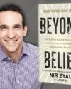 Author Nir Eyal, looking directly at the camera, smiling. Nir has short brown hair and is wearing a blue plaid button down shirt. The book cover of Beyond Belief by Nir Eyal is next to his image with a gray background.
